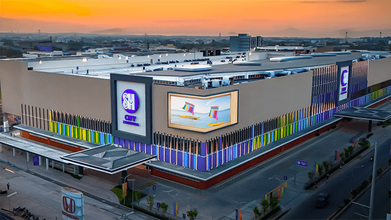SM City Zamboanga illuminated at dusk — the curved outdoor LED wall and building-wide light installations create a landmark presence visible across the city, anchored by CAYIN's centrally managed signage network.